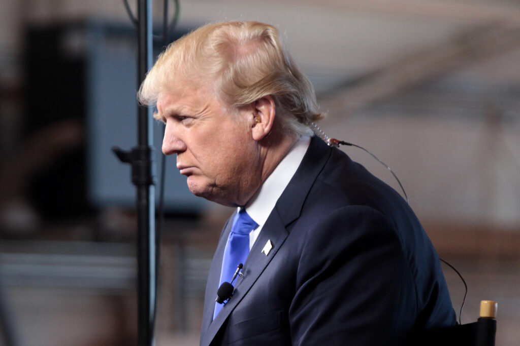 Foto de perfil de Donald Trump. Ele veste um gterno escuro, camisa branca e gravata azul. Sua cabeça está levemente inclinada para a frente