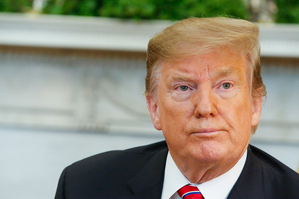 Donald Trump é um homem idoso, com cabelos lisos loiros. Sua pele é alaranjada.